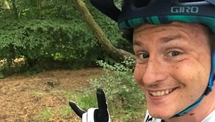 A smiling man wearing a helmet and gloves, posing with his mountain bike in a wooded area