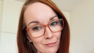 Woman with long auburn hair, wearing glasses and a white top, looks directly at the camera with a slight smile in a well-lit room