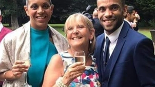 Three smiling people dressed in formal attire hold glasses of champagne at an outdoor event, with a green garden in the background