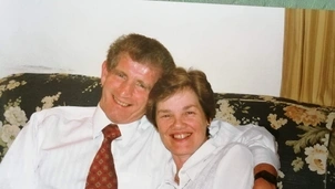 Smiling couple sits closely together on a floral-patterned couch, with the man in a white shirt and red tie wrapping his arm around the woman in a white blouse