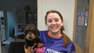 Smiling woman wearing an Asthma + Lung UK t-shirt holds a small black and tan dog in one arm and a glass of bubbly drink in the other, standing in a kitchen