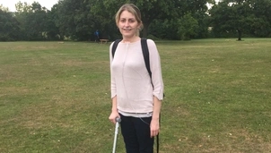 Smiling woman with a backpack stands in a grassy park, using a walking stick and wearing an oxygen tube
