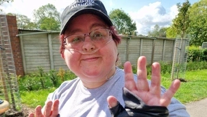 Smiling woman wearing a cap, glasses, and an oxygen tube sits in a wheelchair outdoors, raising gloved hands in a playful gesture