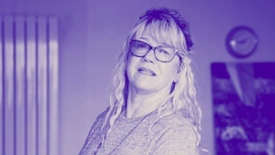 Woman with long wavy hair and glasses, wearing a sweater and necklace, in a room with a clock and radiator