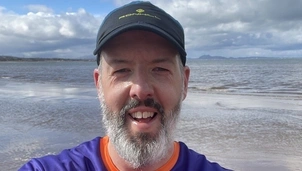 Smiling man with a beard wearing an Asthma + Lung UK t-shirt and a black cap takes a selfie on a beach with the sea and distant hills behind him