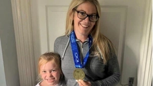 Smiling woman wearing glasses and a grey sports jacket, holding a medal with a blue ribbon, standing beside a young girl with blonde hair and a white t-shirt featuring a pink flamingo