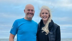 Smiling man in a blue sports shirt and woman in a black jacket stand together outdoors with the ocean and cloudy sky in the background