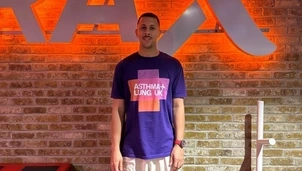 Man wearing an Asthma + Lung UK t-shirt and sportswear stands in front of a brick wall with a large illuminated sign at a gym