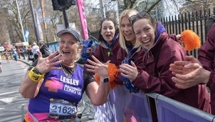 Smiling runner wearing an Asthma + Lung UK vest with race number 21626 poses with hands raised near cheering supporters holding tambourines and pom-poms behind a barrier