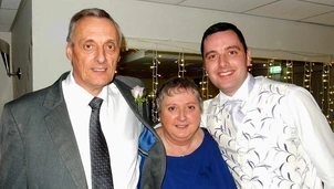 Three smiling people pose at a formal event, with a man in a suit and tie, a woman in a blue dress, and another man in a white patterned waistcoat, against a backdrop of string lights