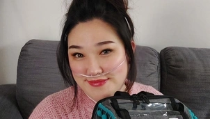 Smiling woman wearing a nasal oxygen tube and a pink sweater sits on a grey couch, holding a decorated medical oxygen bag with a plush toy and badges attached