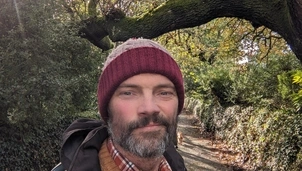 Bearded man wearing a maroon beanie, brown jacket, and backpack takes a selfie on a tree-lined path with sunlight filtering through the branches