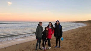 Four people in warm clothing stand together on a sandy beach at sunset, with gentle waves in the background