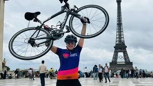 Woman wearing an Asthma + Lung UK cycling jersey and helmet lifts her bike above her head, standing in front of the Eiffel Tower with the Olympic rings