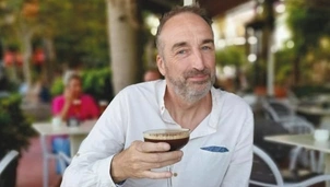 A man in a white shirt holding a coffee drink at an outdoor café, smiling with a relaxed expression