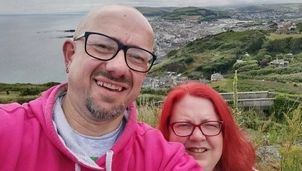 Smiling couple in bright pink hoodies, taking a selfie with a scenic coastal town and green hills in the background