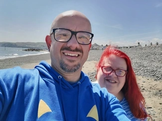 Man wearing a blue hoodie and glasses, smiling alongside a woman with vibrant red hair and glasses, both enjoying a sunny day on a pebbled beach with houses and hills in the background