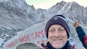 A man smiling in front of a rock marked Everest Base Camp with snowy mountains in the background