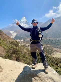 A man standing on a rock with arms raised, wearing hiking gear and a camera, with a snowy mountain peak in the background