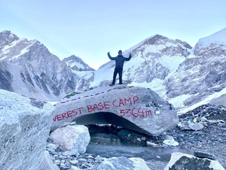 A man standing atop a large rock marked Everest Base Camp 5364m, with snowy mountains in the background
