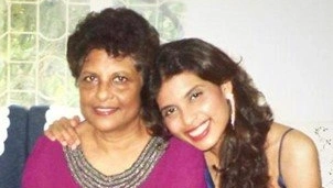Woman in a magenta top sits on a couch, smiling, with a younger woman in a blue dress leaning beside her, wrapping an arm around her shoulder and smiling brightly