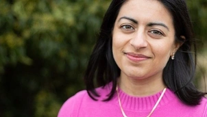 Woman wearing a bright pink sweater and gold necklace, smiling warmly with a blurred green background