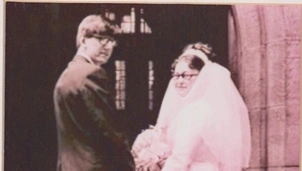 Vintage wedding photo of a bride in a light gown and veil holding a bouquet, and a groom in a dark suit, standing outside a stone building, both wearing glasses