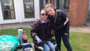 Man in a wheelchair wearing sunglasses sits outside with a young woman hugging him near a picnic table and a brick building