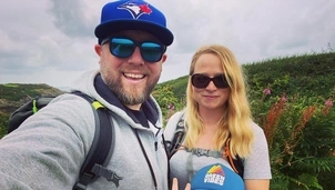 Smiling man in a blue cap and sunglasses takes a selfie with a woman in sunglasses and a young child wearing a hat, posing on a scenic hiking trail with greenery and flowers
