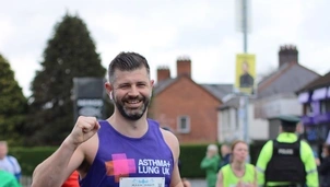 Smiling man wearing an Asthma + Lung UK vest with race number 2821 runs in a marathon, with 'Running for Stevie' printed on his shirt, surrounded by other runners and event staff