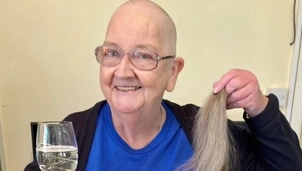 Elderly woman with glasses, smiling while holding a cut ponytail in one hand and a glass of sparkling drink in the other