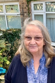 Elderly woman with glasses and long silver hair, smiling gently in a garden setting with greenery and a brick house in the background