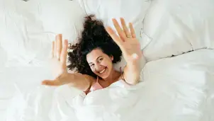 A smiling woman stretching her arms towards the camera from under a white duvet in bed