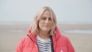 A woman with short blonde hair is stood on a beach looking directly at the camera. She's wearing a red puffa jacket and a blue and white striped t shirt.