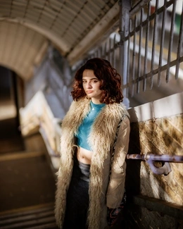 A woman with curly dark hair, in a beige fur-trimmed coat, stands on a staircase, looking thoughtful