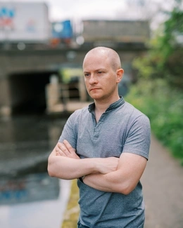 A man with a shaved head, wearing a grey polo shirt, stands with arms crossed by a canal