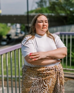 A woman with long hair, wearing a white shirt and animal-print skirt, stands outdoors with arms crossed