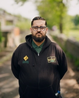 A man with glasses and a beard, wearing a black hoodie, stands outdoors