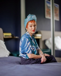A woman with short red hair, wearing a blue headscarf and patterned blouse, sits on a bed
