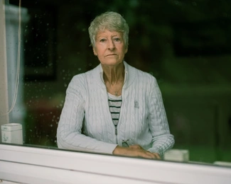 An older woman with short grey hair, wearing a white sweater, looks out thoughtfully through a rain-speckled window