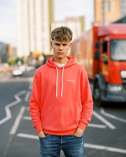 A young man wearing a coral hoodie stands on a city street with a serious expression, hands in his pockets