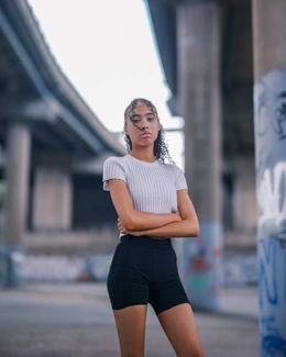 A woman with curly hair, wearing a light grey top and black shorts, stands with arms crossed under an overpass