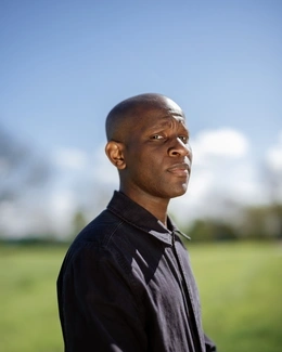 A man with a shaved head, wearing a dark shirt, stands outdoors under a clear blue sky